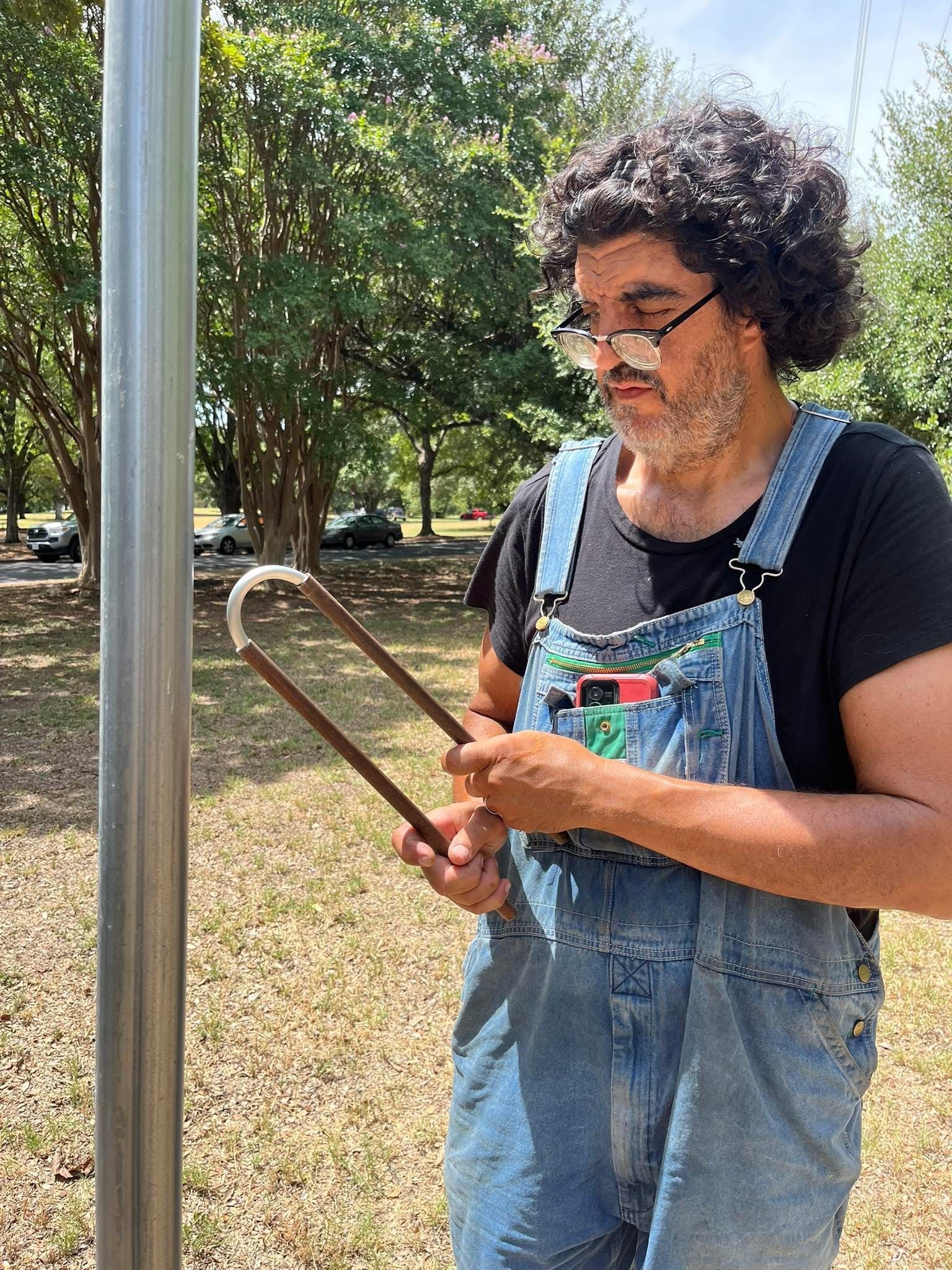 Chalo Colina, in denim overalls and glasses, bending a long rod into a U shape with his hands, outdoors beside a metal pole.