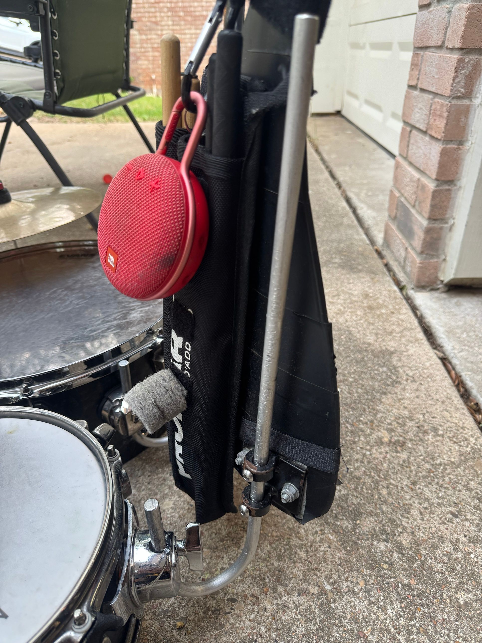 A plain aluminum rod bolted to the harness and bent down into a drum mount on the snare, next to a black stick bag with a red speaker clipped on.